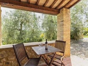 Zona relax all'aperto coperta, con parete in pietra e sedie di legno, affacciata su un giardino alberato.