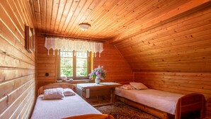 Um quarto de sótão acolhedor, com paredes de madeira, teto inclinado, duas camas de solteiro, uma mesa pequena com um vaso de flores e uma janela com vista para o verde.
