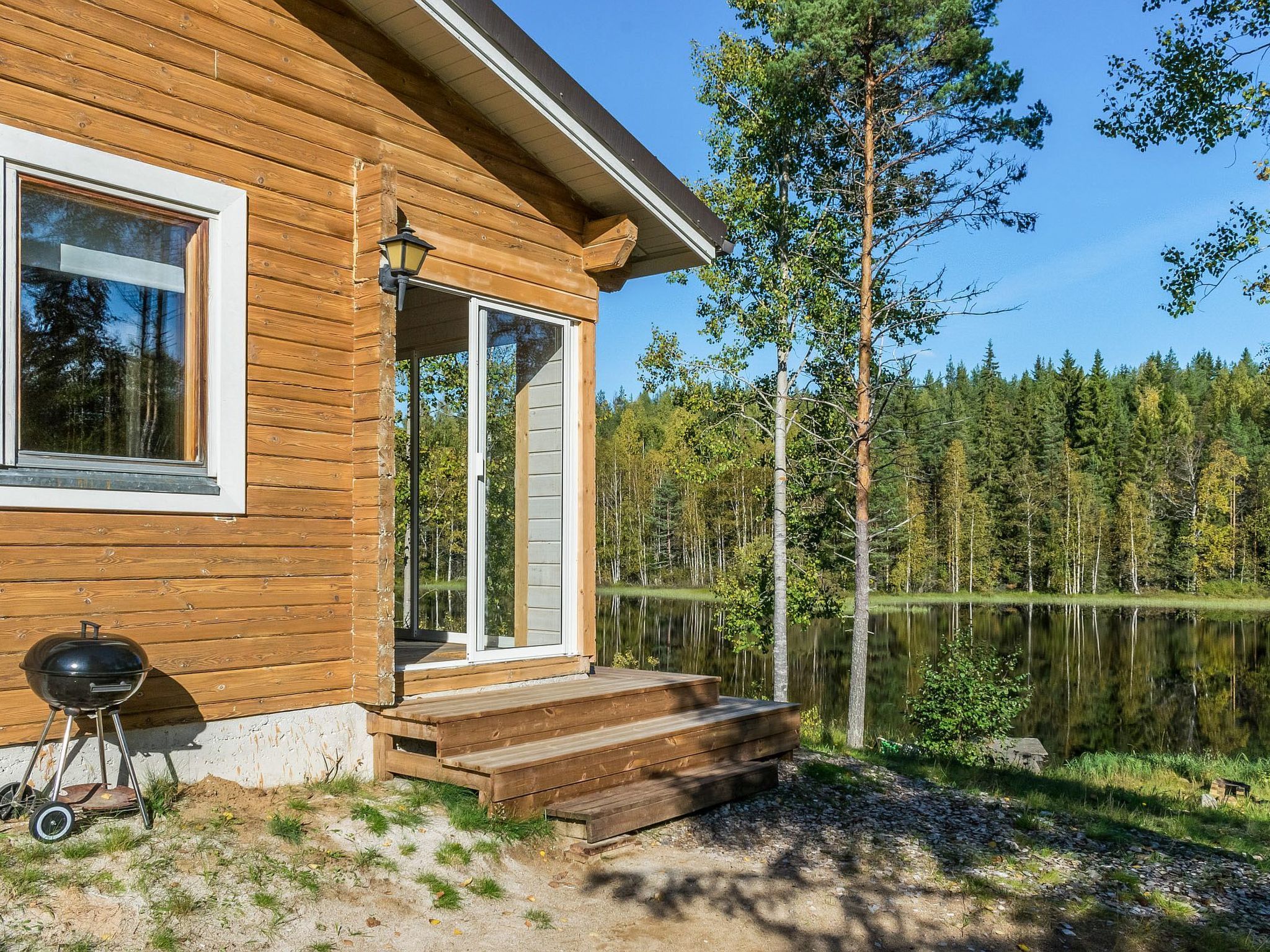 Eine Holzhütte mit Veranda, Grill und Blick auf einen von Bäumen umgebenen See.