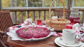 Una mesa de madera con un pastel de vidrio sobre un mantel de encaje, una cesta de mimbre, un jarro de bebida roja y una taza blanca en una terraza de madera con vistas a árboles.