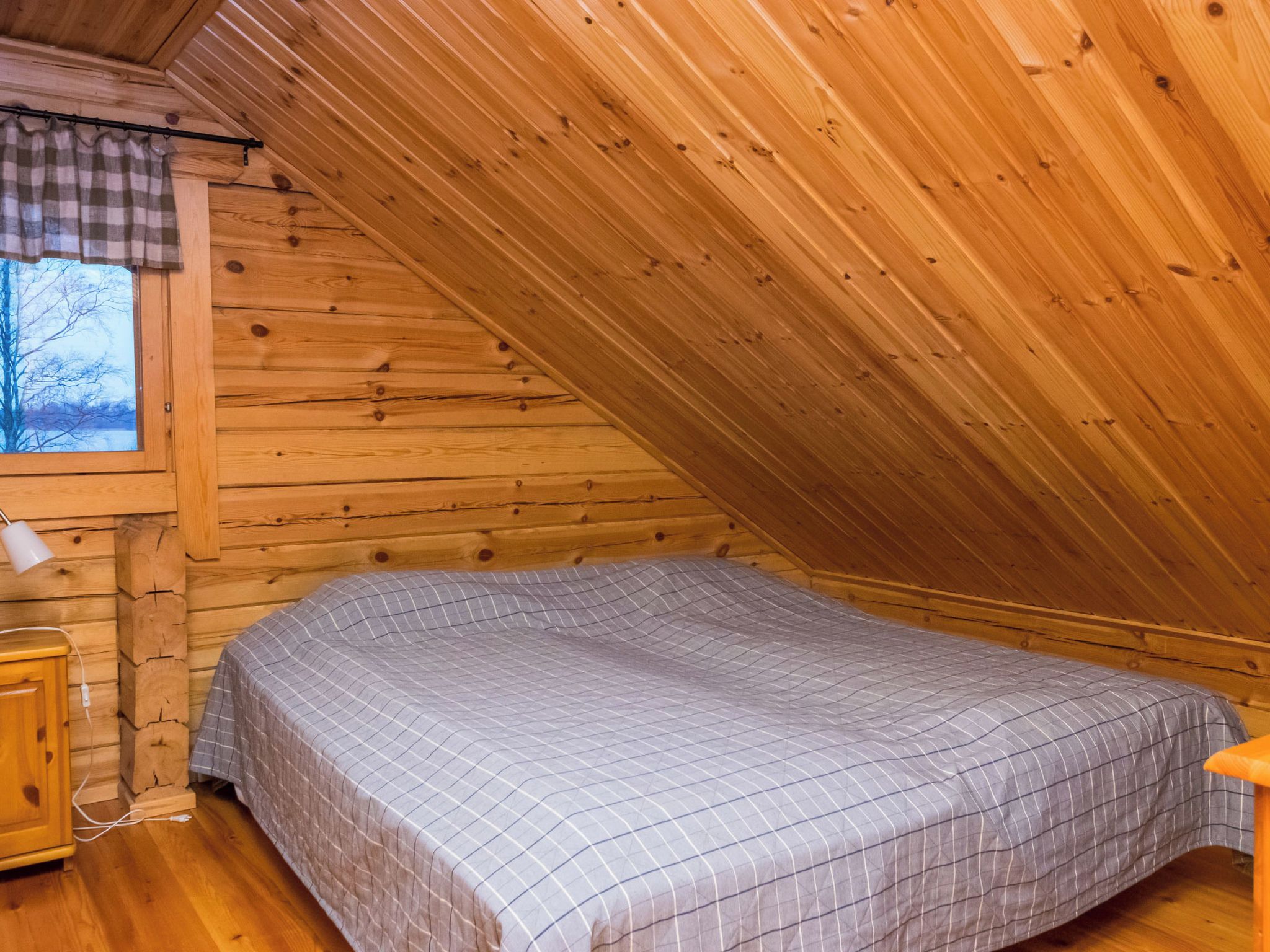 Ein kleiner, gemütlicher Raum mit schräger Decke, einem Einzelbett mit karierter Bettdecke, einem Holznachttisch und einem Fenster mit Blick auf kahle Bäume.
