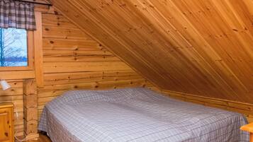 Ein kleiner, gemütlicher Raum mit schräger Decke, einem Einzelbett mit karierter Bettdecke, einem Holznachttisch und einem Fenster mit Blick auf kahle Bäume.