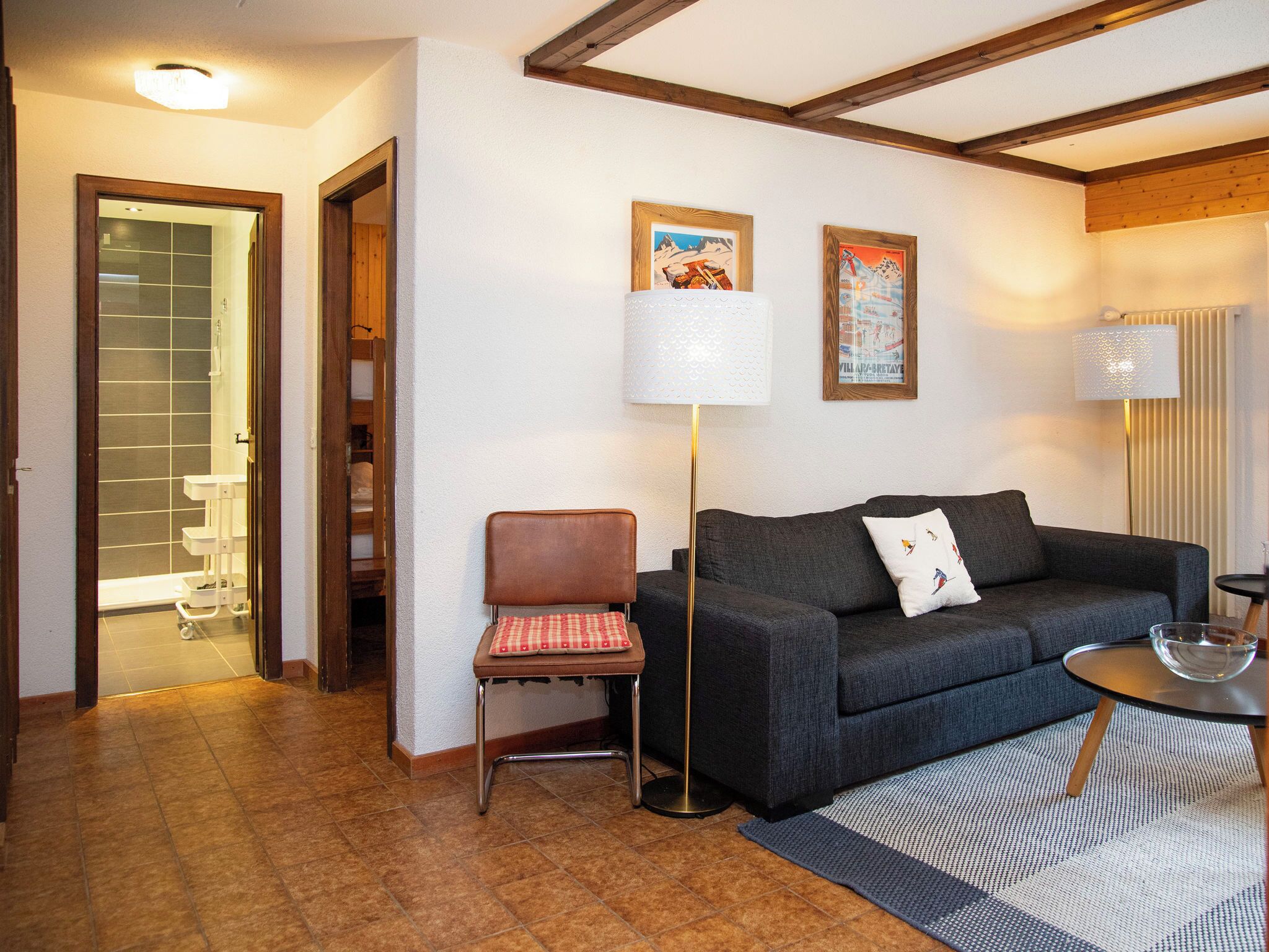 A living room with a dark grey sofa, a small round table, a floor lamp, and a chair with a red and white checkered cushion. There are two framed pictures on the wall, a wooden ceiling beam, and a tiled floor. - Opale 4 by Interhome (Villars)