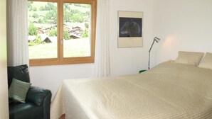 Ein Schlafzimmer mit Bett, Stuhl, Schreibtischlampe und Fenster mit Blick auf Häuser und Grünflächen.