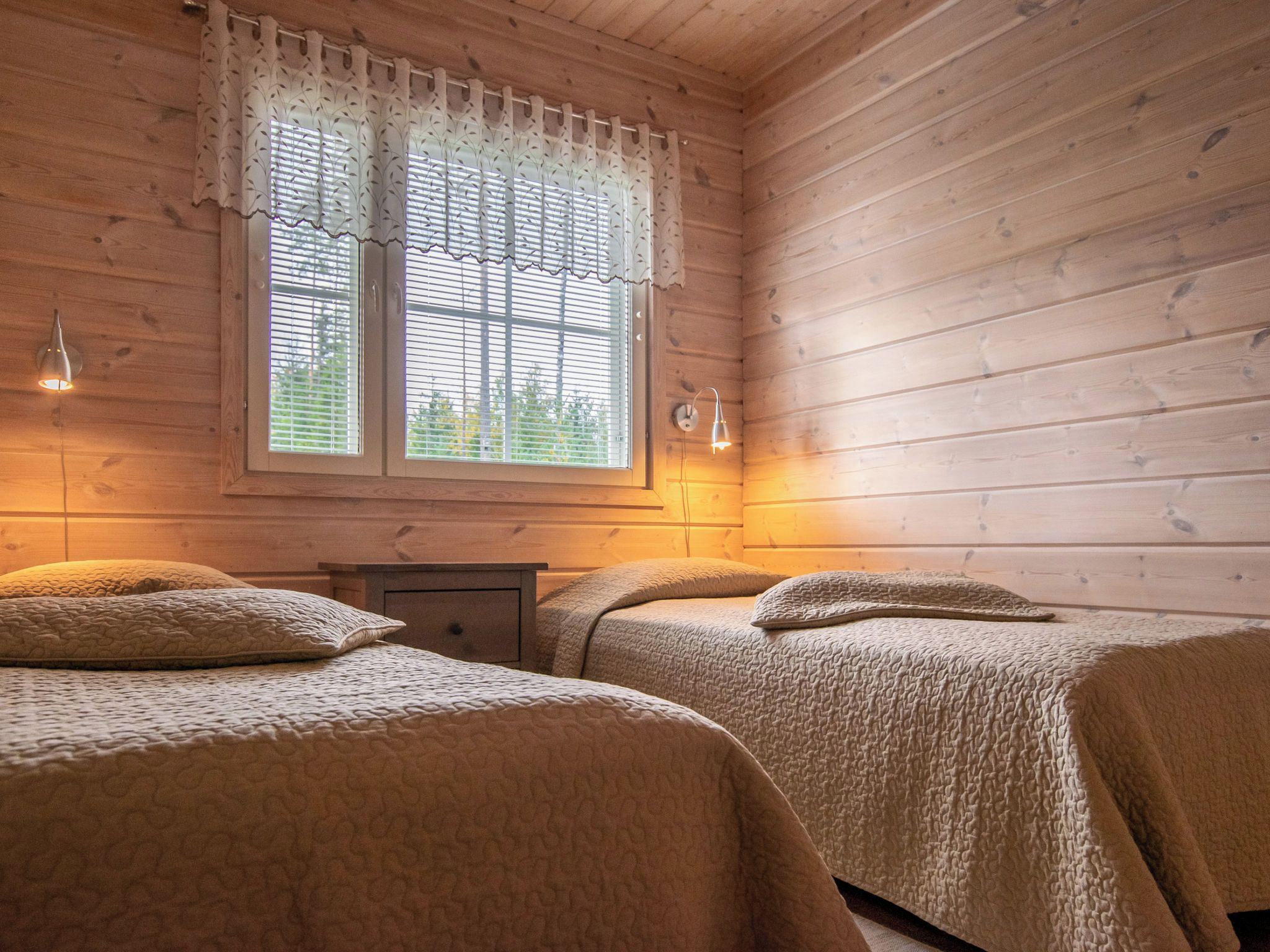 Ein Zimmer mit Holzboden, zwei Betten, einem Nachttisch und einem Fenster mit Vorhängen.