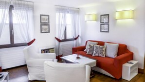 Una sala de estar moderna con un sofá rojo, sillones blancos y una mesita. Hay dos ventanas con cortinas translúcidas y lámparas fijadas a la pared.