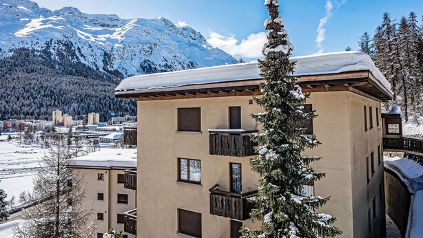 Un edificio con varios balcones y ventanas, rodeado de árboles nevados y montañas al fondo.