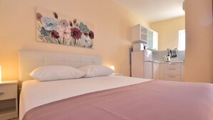 Ein Hotelzimmer mit Bett, Nachttisch, einem Blumenbild und einer Küchenzeile mit Schränken und einem Kühlschrank.