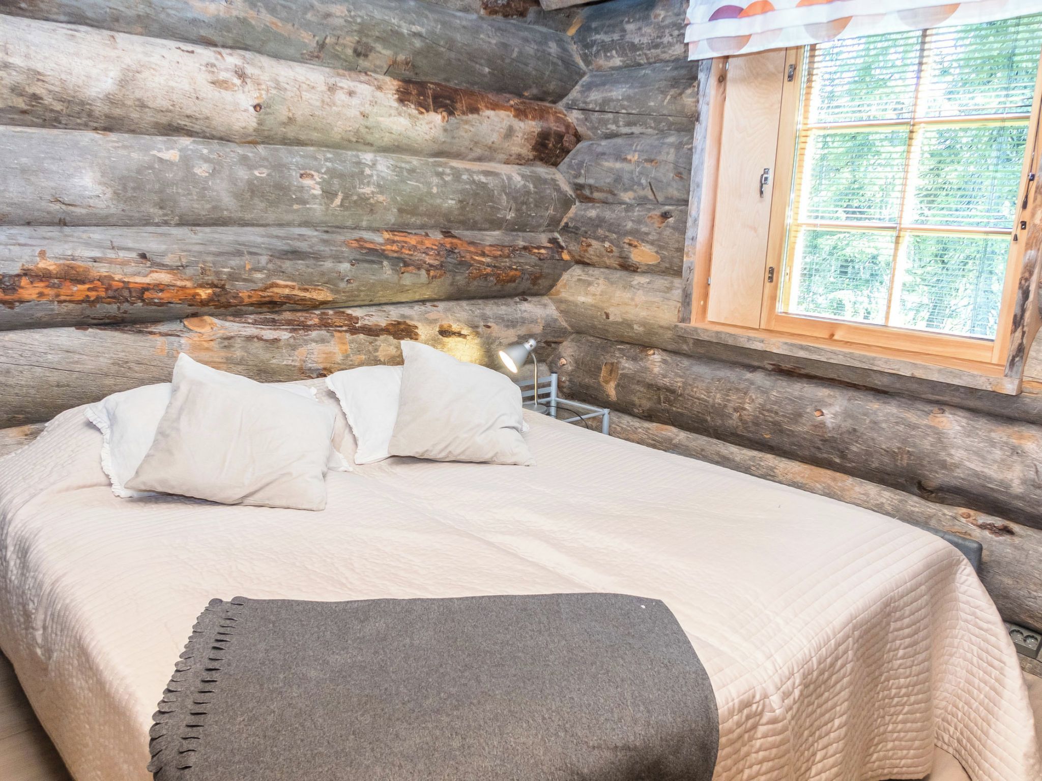 Une chambre aménagée dans un chalet en rondins, avec un lit, des oreillers, une lampe et une fenêtre donnant sur un paysage agréable.