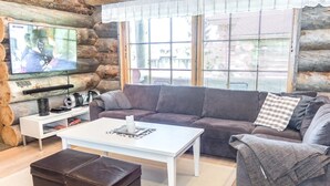 Un salon chaleureux avec un mur en pierre, une grande fenêtre avec des stores, un canapé gris, une table basse blanche et un téléviseur posé sur un meuble en bois.