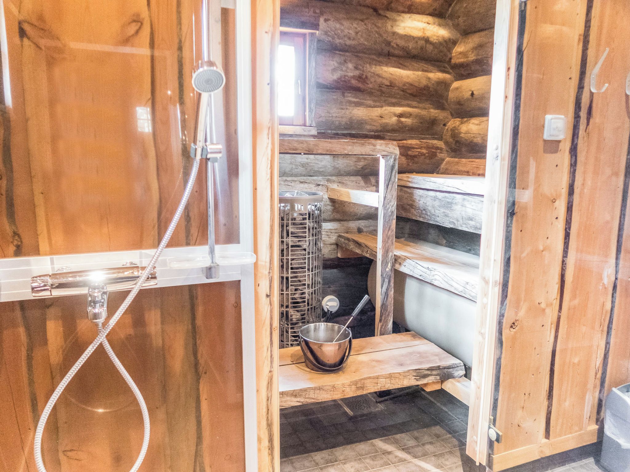 A wooden cabin with a shower, a metal bucket, and a window.