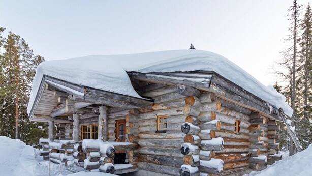 Une cabane en rondins dont le toit et les murs extérieurs sont recouverts de neige, entourée de neige et d’arbres.
