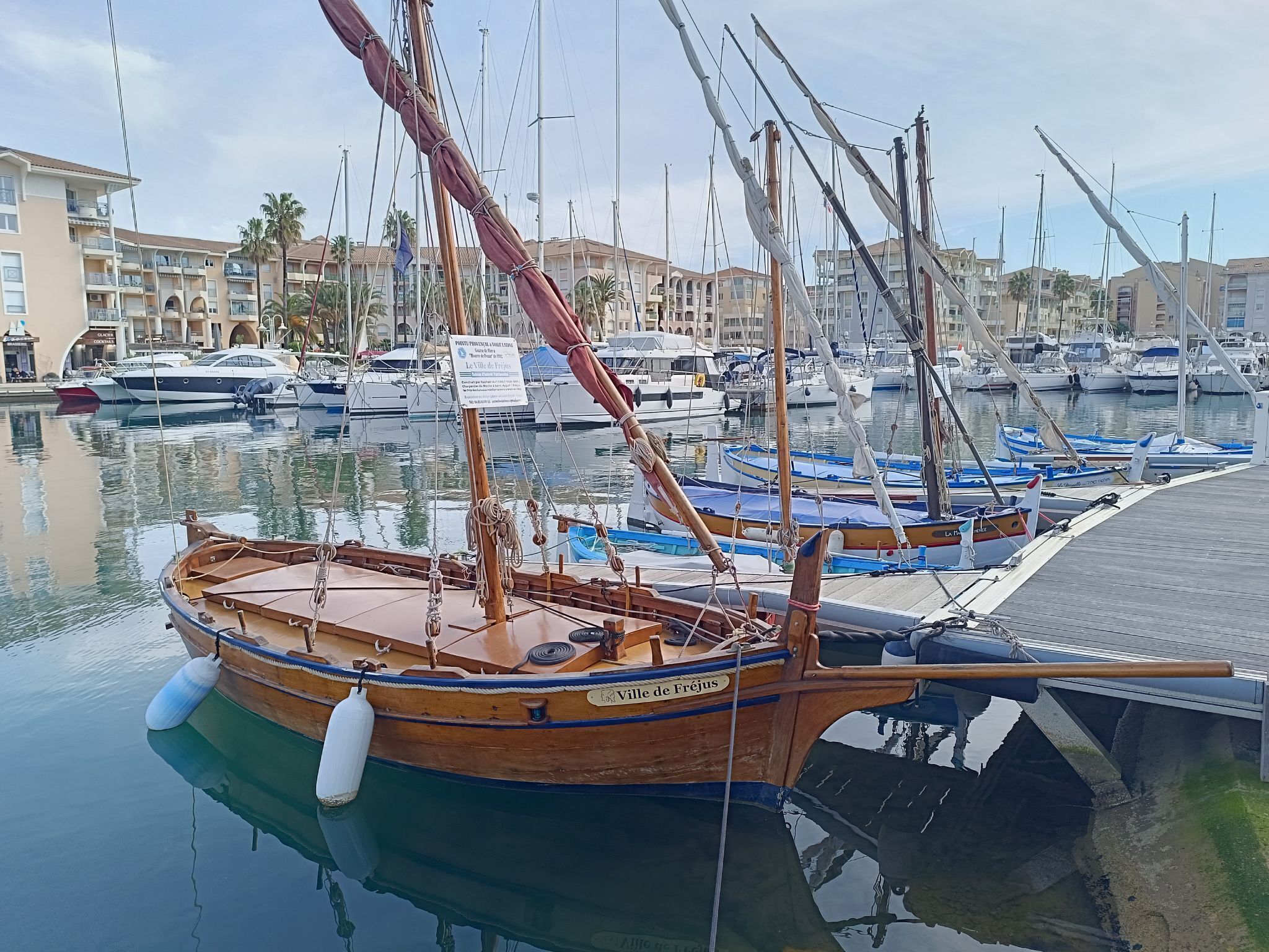 Un port de plaisance avec des bateaux amarrés, un bateau en bois nommé « Ville de Fréjus », et un panneau indiquant « Pointe Peronne à Virel ».