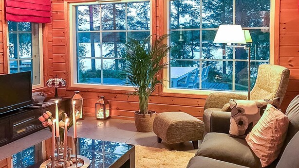 Una sala de estar acogedora con televisor, una mesa de centro de vidrio, un sofá y un sillón. Hay ventanas grandes que ofrecen vistas a árboles y un edificio.