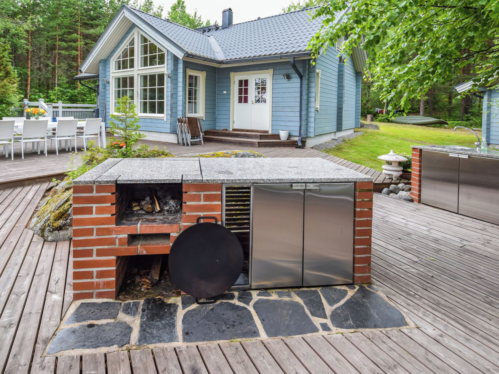A house with a blue exterior, a brick barbecue area with a stone surround, and a deck with outdoor furniture.