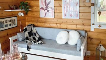 Una habitación acogedora con un sofá gris, cojines blancos, una manta de crochet, cuadros en la pared, una planta y una mesita de mimbre con vasos.