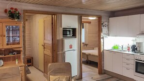 A compact kitchen with a microwave, refrigerator, and sink. There is a wooden dining table with a chair. The room has a wooden floor and a colorful rug.