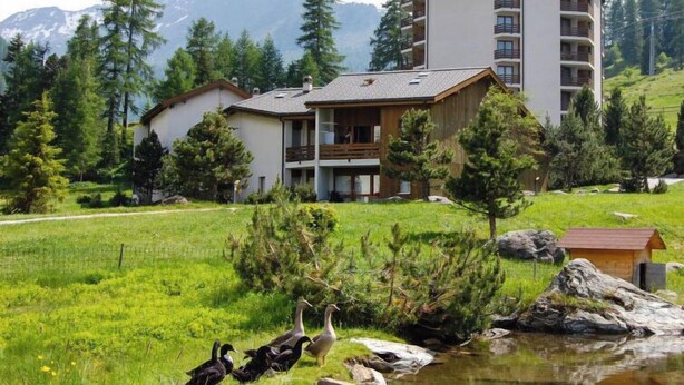 A modern hotel with a large building and a smaller annex, surrounded by lush greenery and a pond with ducks. - Mont-Fort 51 by Interhome (Siviez-Nendaz)