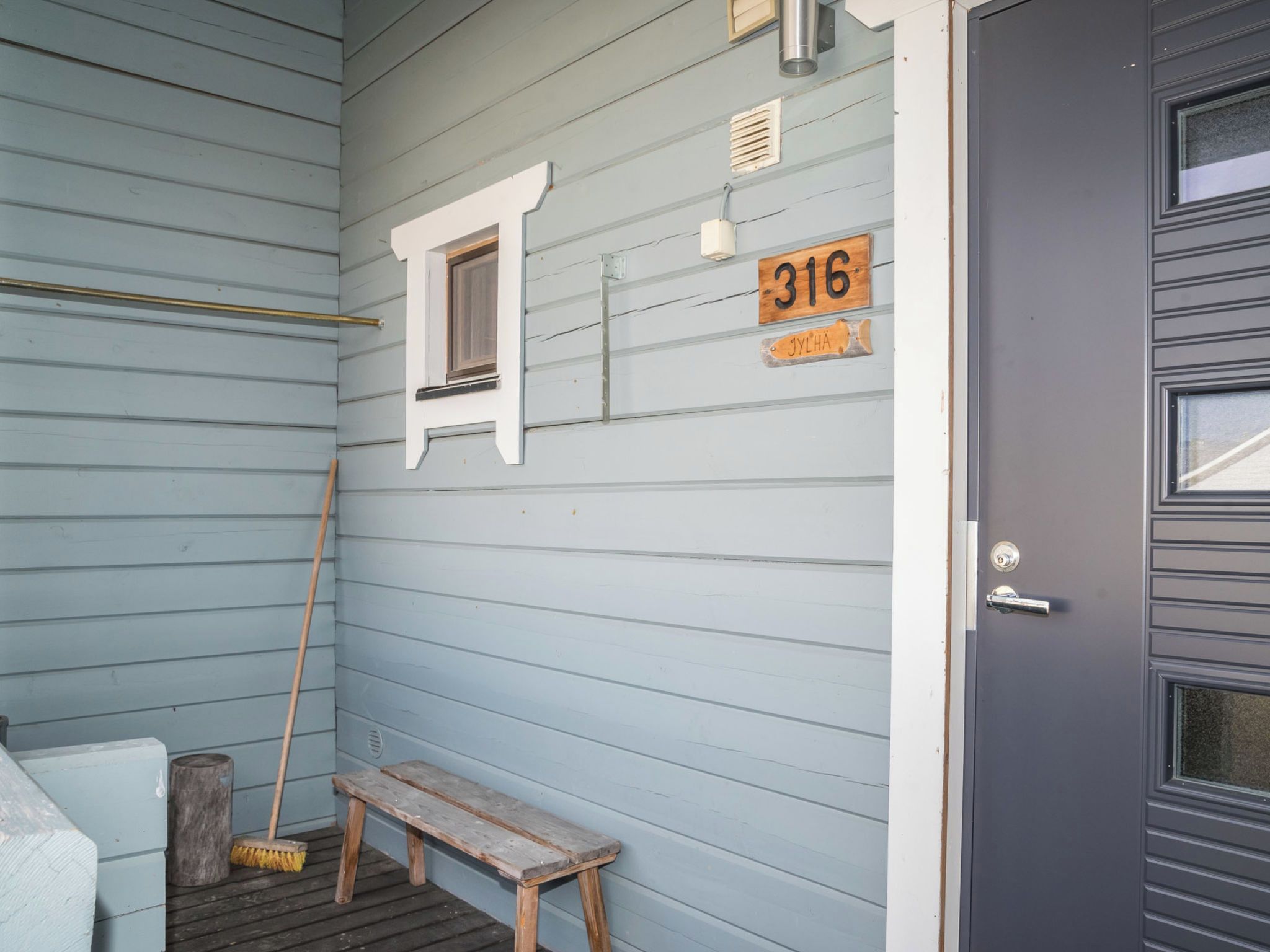 A wooden bench, a broom, a window, and a door with a number plate.