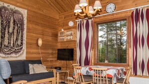 Un acogedor interior de cabaña con detalles de madera, un sofá, una mesa de comedor con sillas, un televisor y un ventanal con cortinas.