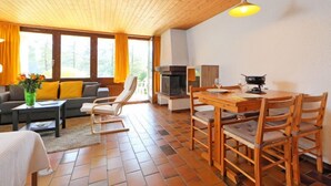Ein gemütlicher Wohnbereich mit einer Couch, einem Esstisch mit Stühlen, einem Kamin und einem großen Fenster mit Vorhängen.