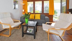 Ein gemütliches Wohnzimmer mit Holzboden, zwei weißen Sesseln, einem grauen Sofa, einem Couchtisch mit Blumenvase und großen Fenstern mit gelben Vorhängen.