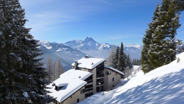Ein modernes Skiresort-Gebäude mit mehreren Balkonen, umgeben von schneebedeckten Bäumen und Bergen im Hintergrund.