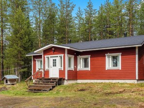 A red wooden cabin with a front porch and a chimney, surrounded by a forest.