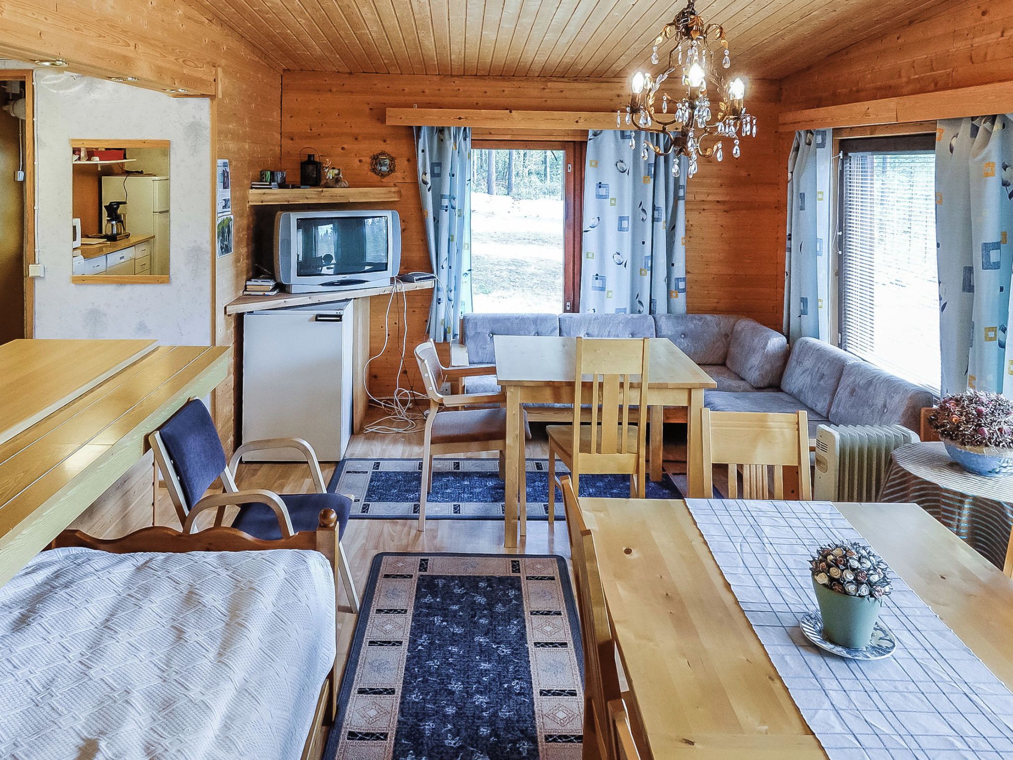 Un accogliente interno di una baita con soffitto in legno, zona pranzo, divano, televisore e un piccolo frigorifero.