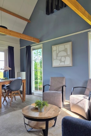 Un soggiorno moderno con soffitto alto, un'area pranzo con tavolo e sedie in legno e un'ampia finestra con tende.