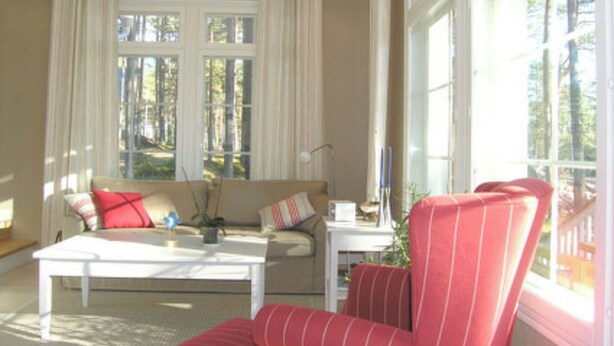 Ein Wohnzimmer mit Sofa, Sessel und Couchtisch. Große Fenster mit Vorhängen, eine Lampe und eine Vase mit Blumen sind ebenfalls zu sehen.