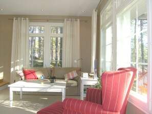 Una sala de estar con sofá, sillón y mesa de centro. Hay ventanas grandes con cortinas, una lámpara y un florero con flores.