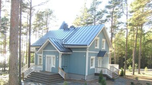 Ein zweistöckiges Haus mit einem Metall-Dach, einer Veranda mit Stufen und einem kleinen Vorgarten mit Steinen und jungen Bäumen.