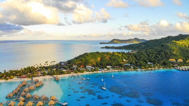 An aerial view of a tropical resort with overwater bungalows, a beach, and clear blue waters.