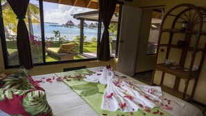 A hotel room with a bed, a view of a beach and overwater bungalows, and a wooden shelf with toiletries.