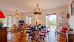 Un comedor con una mesa dispuesta para cuatro personas, rodeada de sillas de mimbre, una lámpara roja y un colgador de luces.