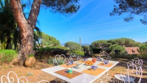 Un área de comedor al aire libre con una mesa y sillas de metal blanco, rodeada de árboles y un cielo despejado.