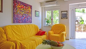 A room with a yellow sofa, a chair, a table with a fruit bowl, a painting, and a window with a view of greenery. - Apartment Andrea by Interhome (Opatija/Bregi)