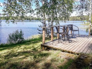 Une terrasse en bois avec une table et des chaises, donnant sur un lac paisible, entourée d’arbres et d’herbe.