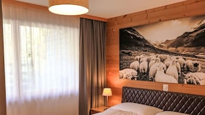 A hotel room with a bed, a nightstand, a lamp, a window with curtains, and a wall-mounted painting of sheep.