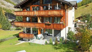A multi-story wooden chalet with balconies, large windows, and a mountainous backdrop.