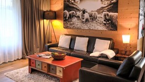 A modern living room with a wooden wall, a large painting of sheep, a leather sofa, a coffee table with a fruit bowl, and a floor lamp.