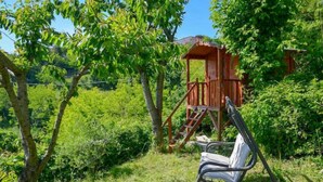 A wooden playhouse with a ladder, surrounded by lush greenery and a metal swing chair.