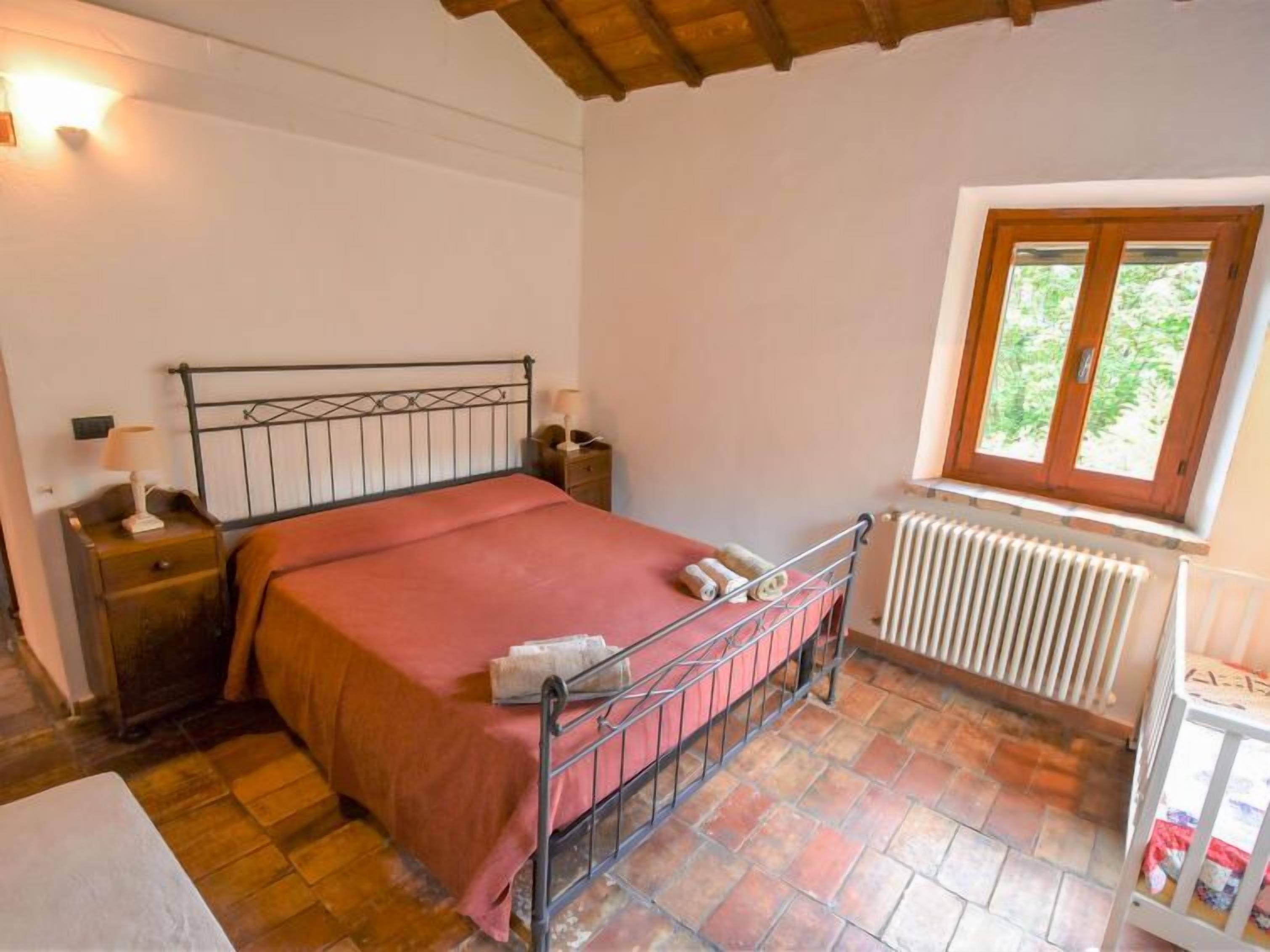 A bedroom with a metal bed frame, a wooden headboard, a red bedspread, a nightstand with a lamp, a window with a view of greenery, and a radiator.