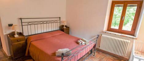 A bedroom with a metal bed frame, a wooden headboard, a red bedspread, a nightstand with a lamp, a window with a view of greenery, and a radiator.