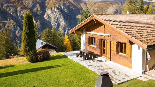 A chalet with a stone foundation, wooden exterior, and a tiled roof. It features a patio with a table and benches, surrounded by a well-maintained lawn and a tall evergreen tree. - Als man zählte 1989 by Interhome (St Niklaus)