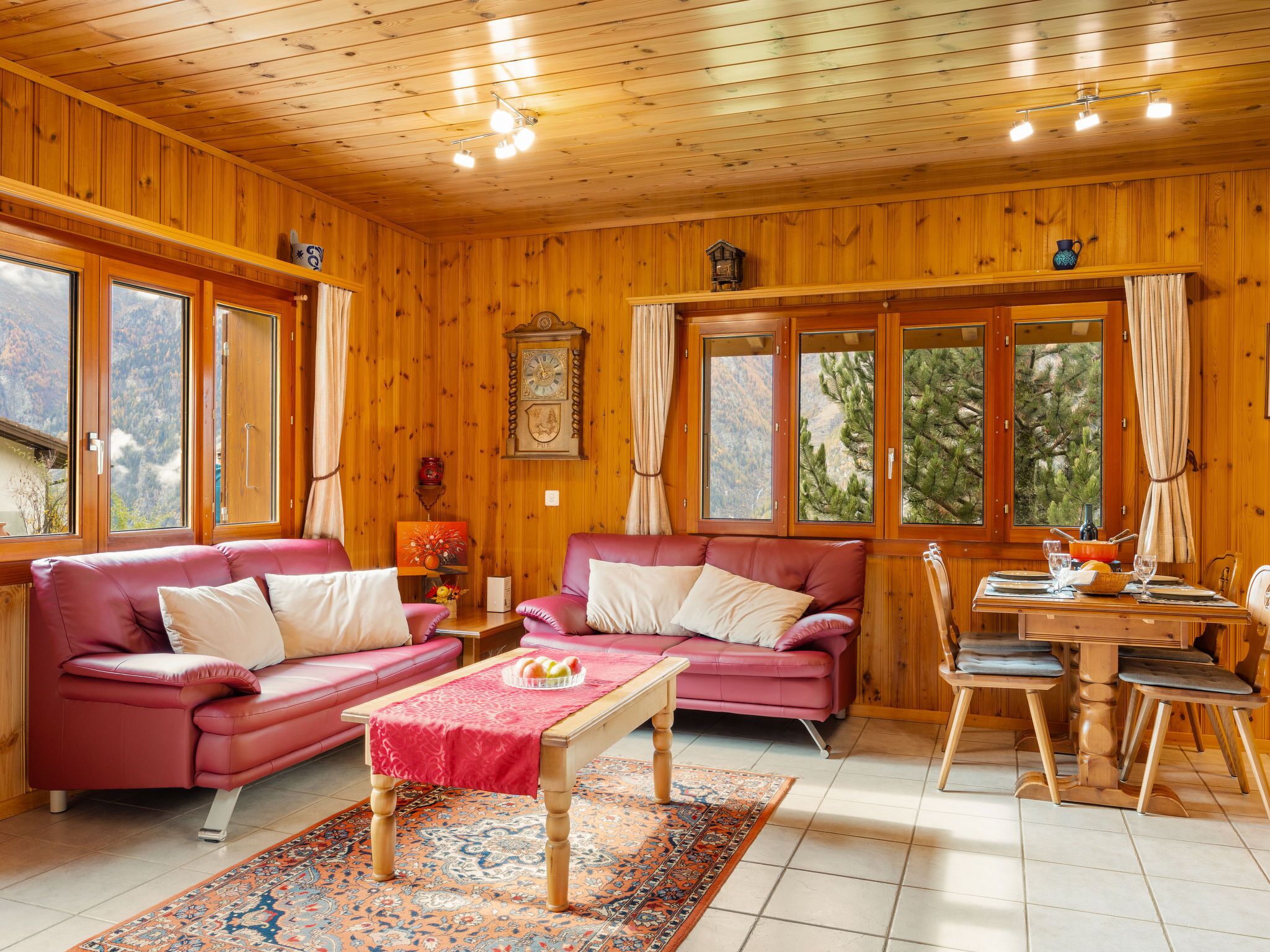 A cozy wooden cabin with two red leather sofas, a wooden dining table, and large windows offering mountain views.