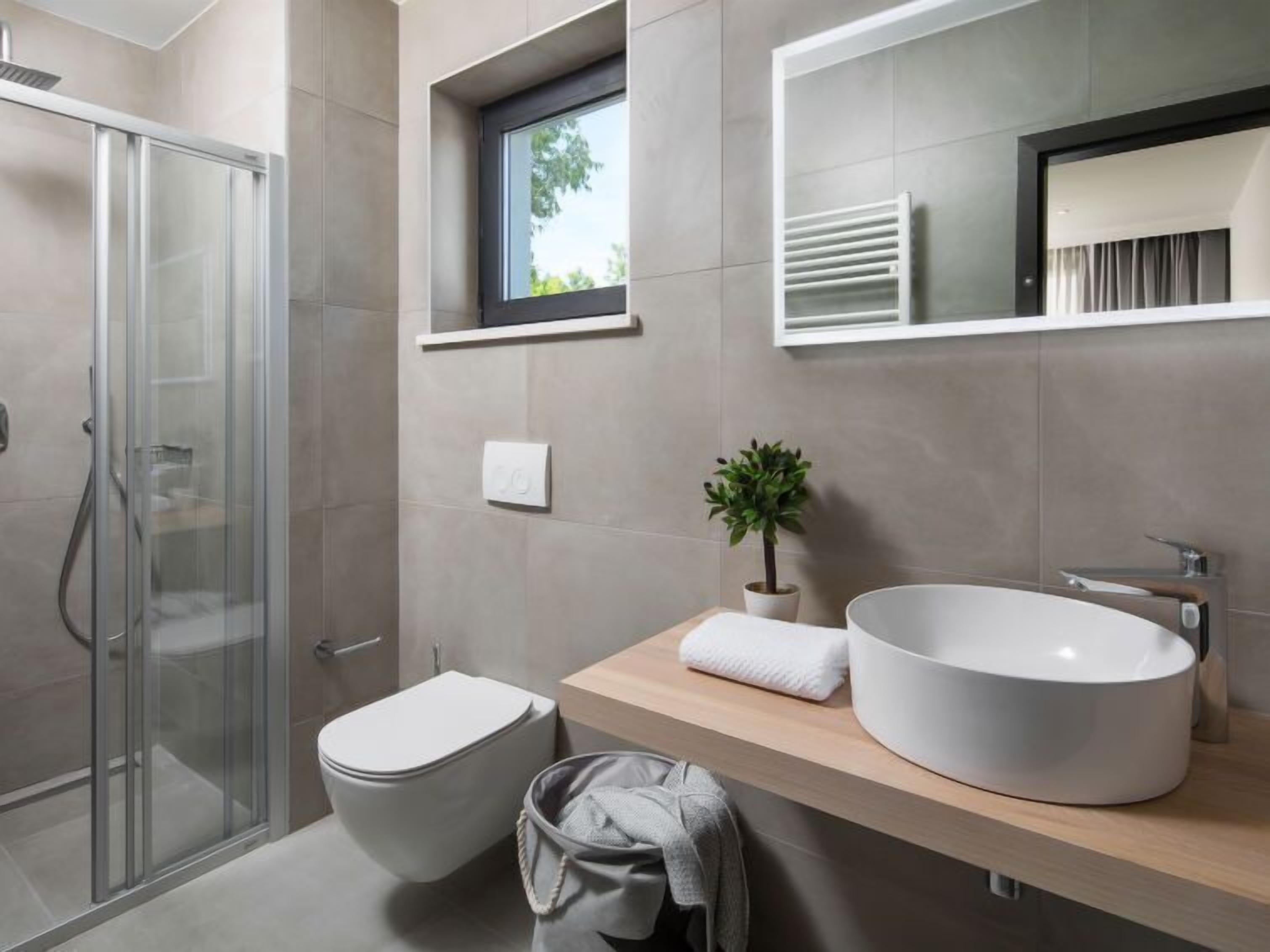A modern bathroom with a glass-enclosed shower, a toilet, a sink with a large basin, a small window, and a potted plant.