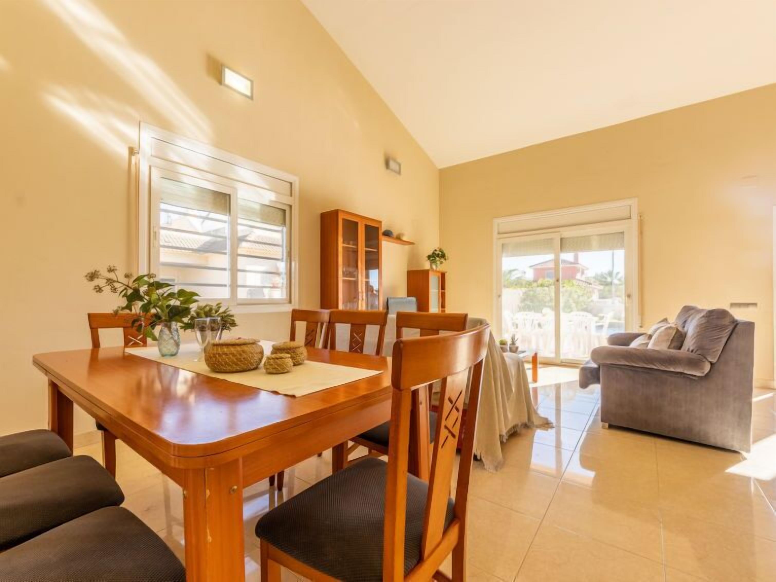 A dining area with a wooden table and chairs, a cabinet, and a sofa in a well-lit room with large windows.
