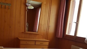 A room with wooden paneling, a wooden dresser, a mirror, two beds with white bedding, and a window with red checkered curtains.